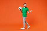 Full length portrait excited man football fan in green t-shirt cheer up support favorite team with soccer ball pointing index finger aside isolated on orange background. People sport leisure concept.