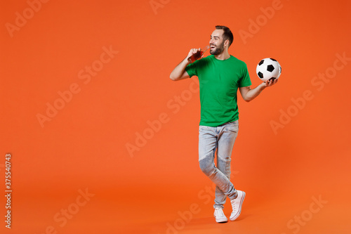 Fototapeta Naklejka Na Ścianę i Meble -  Full length portrait cheerful young man football fan in green t-shirt cheer up support favorite team with soccer ball hold beer bottle isolated on orange background. People sport leisure concept.