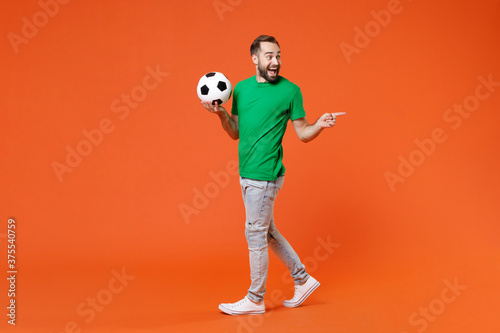 Fototapeta Naklejka Na Ścianę i Meble -  Full length portrait excited man football fan in green t-shirt cheer up support favorite team with soccer ball pointing index finger aside isolated on orange background. People sport leisure concept.