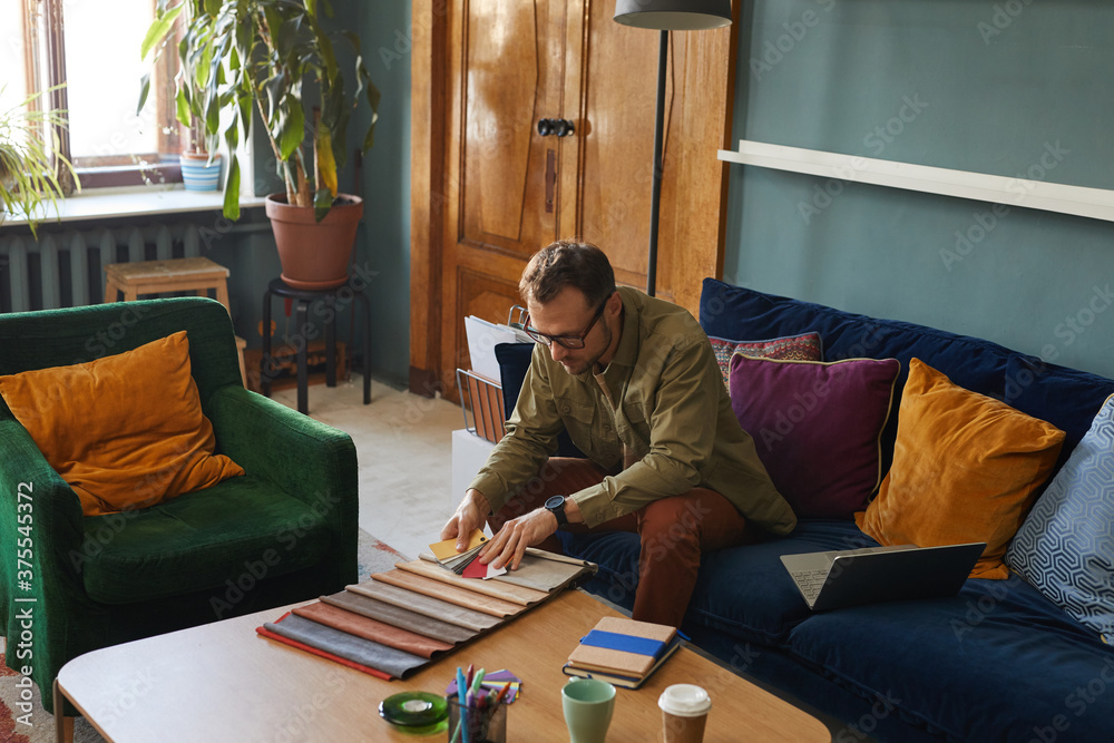 Fototapeta premium Mature designer in eyeglasses sitting on sofa in front of the table and working with fabric samples at modern office