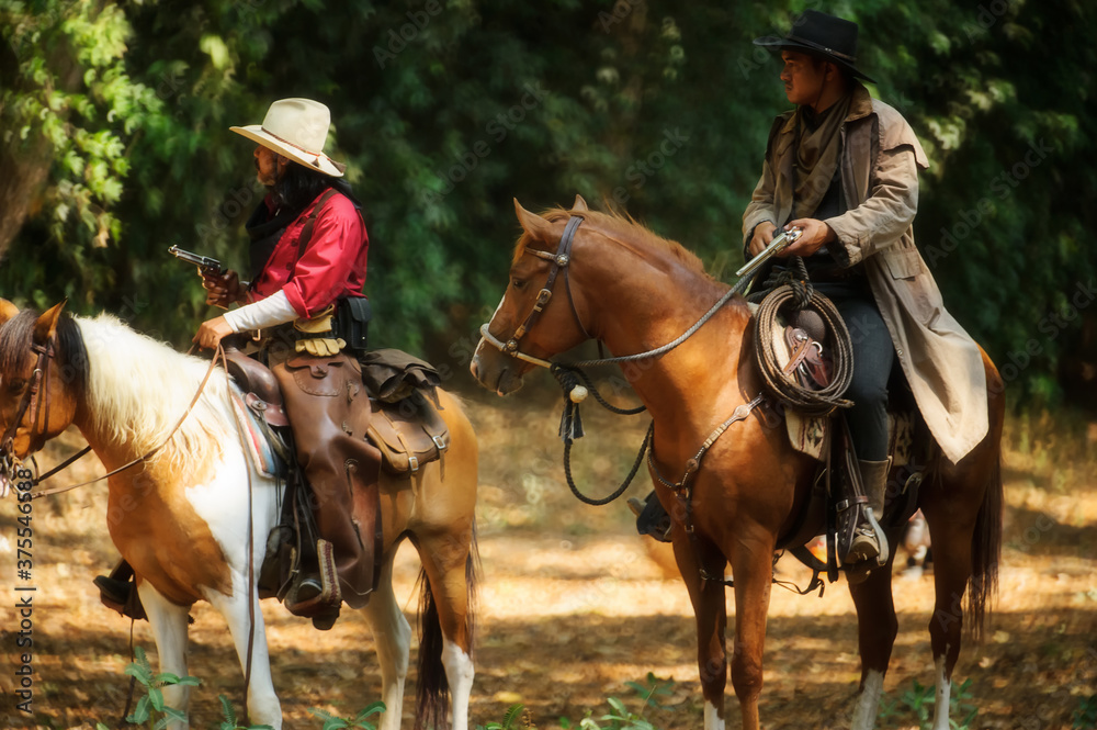 Two cowboy is on the horse, standing in the forest and holding a gun.
