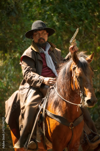 The cowboy is on the horse, standing in the forest and holding a gun.