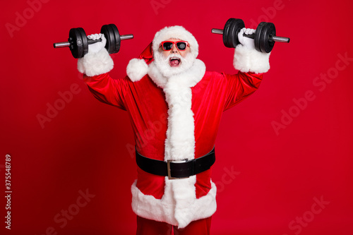 Photo of pensioner old mad man white beard lift dumbbells open mouth screaming heavy weight ignoring muscle pain wear santa x-mas costume sunglass cap isolated red color background