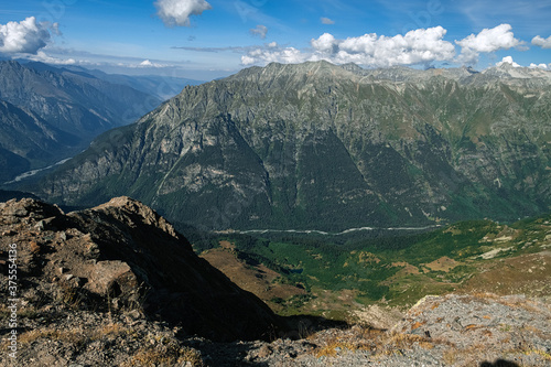 Large-scale mountain landscape from the height of the mountains. A small river in the distance and a lake. Cloudy blue sky.