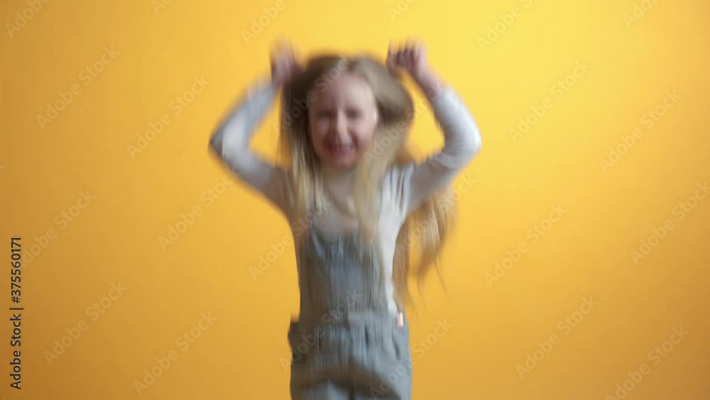 Portrait of happy joyful cute blonde little girl, bounces up. Winning win concept. Yellow background. Girl in denim overalls casual wear. Portrait of happy joyful cute blonde little girl, bounces up. Winning win concept. Yellow background. Girl in denim overalls casual wear.