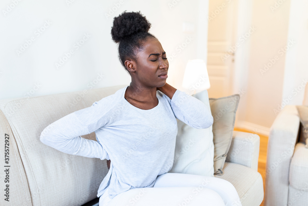 Young Black woman suffering from backache at home. Portrait of a young girl sitting on the couch at home with a headache and back pain. Beautiful woman Having Spinal Or Kidney Pain Young Black woman suffering from backache at home. Portrait of a young girl sitting on the couch at home with a headache and back pain. Beautiful woman Having Spinal Or Kidney Pain