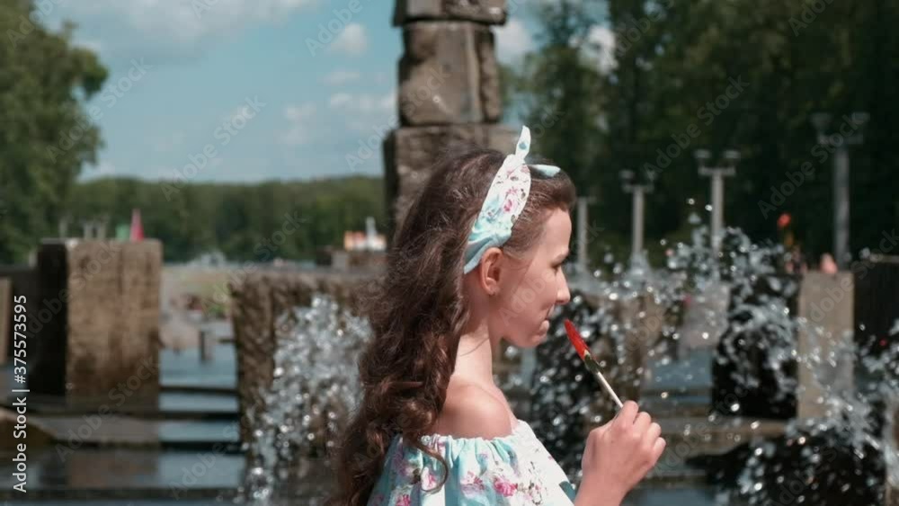 A beautiful girl in a full-length dress near the fountain. There is a sweet candy on a stick. Long beautiful hair. Rest on a day off in the park outside. Slow motion. A beautiful girl in a full-length dress near the fountain. There is a sweet candy on a stick. Long beautiful hair. Rest on a day off in the park outside. Slow motion.