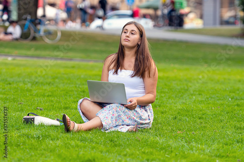 a young woman sitting on a lawn in a city park and working on a laptop, distance learning and working concept