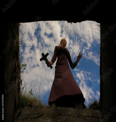 black widow Holding a stone cross in hand. Halloween woman in gothic dress crying on a tomb. blue cloudy sky background.