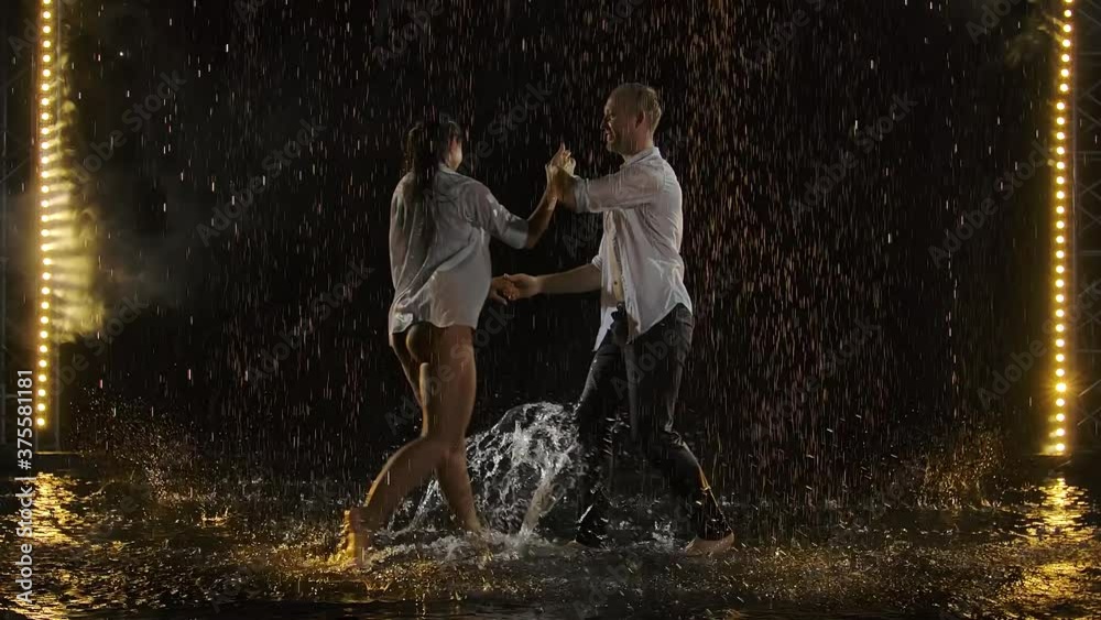 Skillful dancers dance salsa in a dark room under the concert lights and smoke. The sensual couple performs an artistic and emotional dance among the raindrops and many splashes. Slow motion.