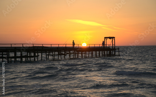 Two fishermen silhouette on a dock in Mediterranean Sea against a background of golden horizon at sunset time