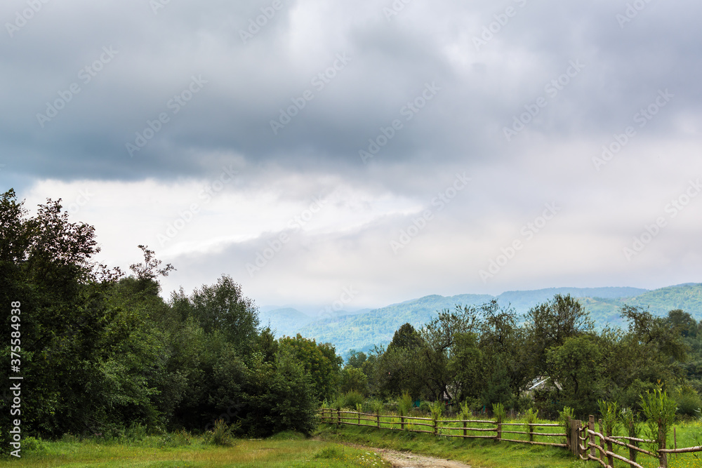 Nature in the village. Trees, mountains in the background in the fog. Green and blue colors. The fence and the road are brown. Calm and rest. Clouds. Gloomy sky. Nature in the village. Trees, mountains in the background in the fog. Green and blue colors. The fence and the road are brown. Calm and rest. Clouds. Gloomy sky.