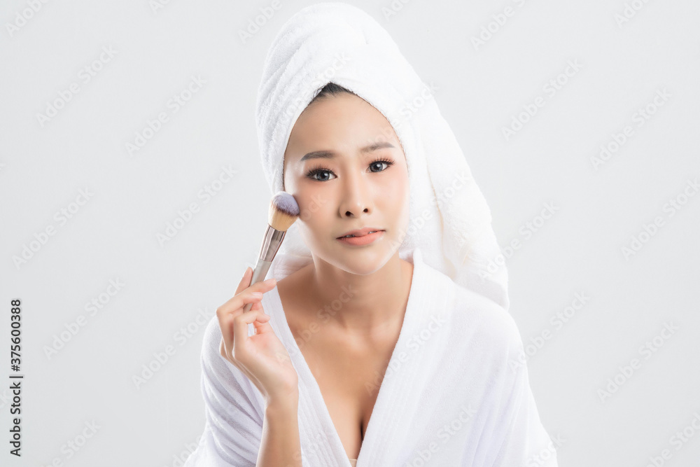 Beautiful young woman wearing bathrobe with towel with towel on head is using a makeup brush makeup her after bathing, at studio shot isolated on white background. Beautiful young woman wearing bathrobe with towel with towel on head is using a makeup brush makeup her after bathing, at studio shot isolated on white background.