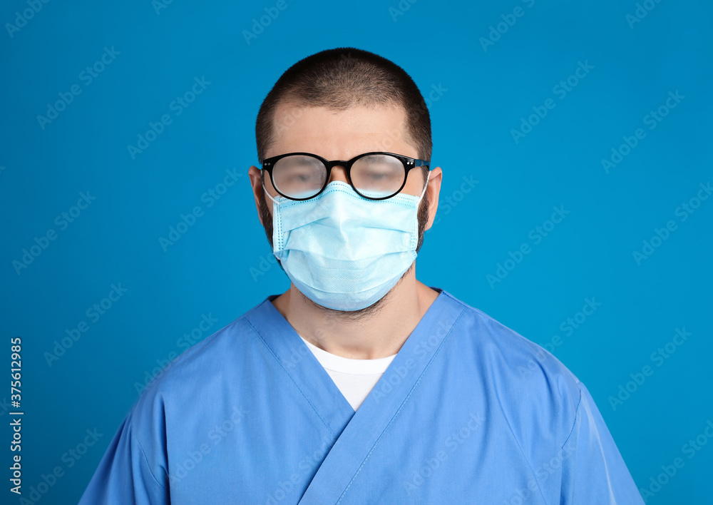 Doctor with foggy glasses caused by wearing disposable mask on blue background. Protective measure during coronavirus pandemic Doctor with foggy glasses caused by wearing disposable mask on blue background. Protective measure during coronavirus pandemic