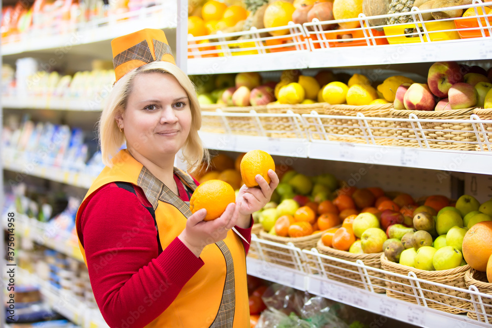 The saleswoman in the supermarket near the shelf with fruit.The seller in a supermarket with three hands