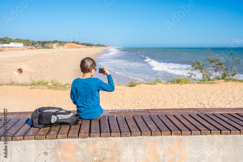 Back view of boy photographs or shoots video of the sea on a mobile phone. Beautiful day sunny outdoors background.