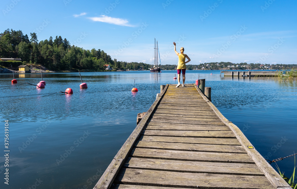 Young woman stands on a wooden pier on the sea bay and waves to moving sailing yacht. Slender beautiful Girl in shorts. Blond Female on shore in the Baltic sea bay in Scandinavia. Panorama.