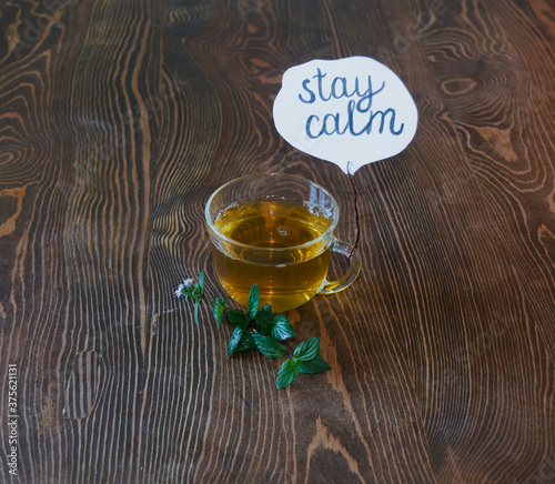 A mug with a decoction of herbs and fresh sprigs of mint on a wooden table. Close-up. There is a plate attached to the cup of tea, stay calm. The quote is written in ink on paper. Morning mood.