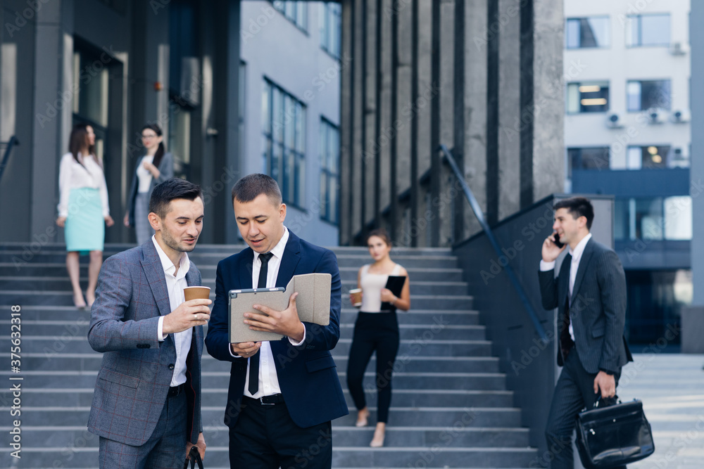 Formal dressed Businessmen speaking and walking outside office building. Businessman discussing something and disputing with Caucasian businessman with tablet computer in hands Formal dressed Businessmen speaking and walking outside office building. Businessman discussing something and disputing with Caucasian businessman with tablet computer in hands