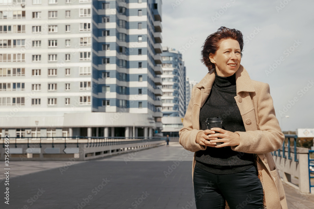 Fototapeta premium Woman in beige coat walks along the road along modern high-rise buildings