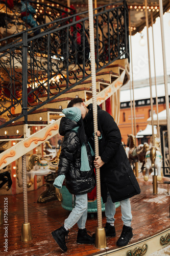 Young couple in warm clothes embracing and kissing outdoors on the carousel in amusement park. Winter holidays.