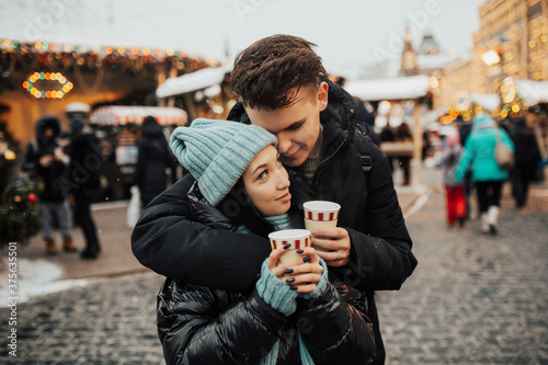Having fun together at a Christmas market. Young cheerful couple is having a walk with hot drinks, enjoying holiday time. They are dressed warm.