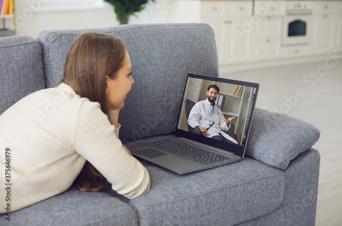 A young woman lies on the couch at home and makes a video call to the doctor online using a laptop. Videoconference with a patient.