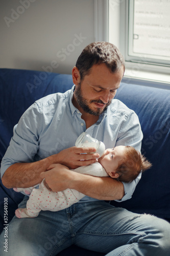 Middle age Caucasian father feeding newborn baby from bottle with milk. Man parent holding rocking child on his hands. Authentic lifestyle candid real moments. Fathers Day holiday.