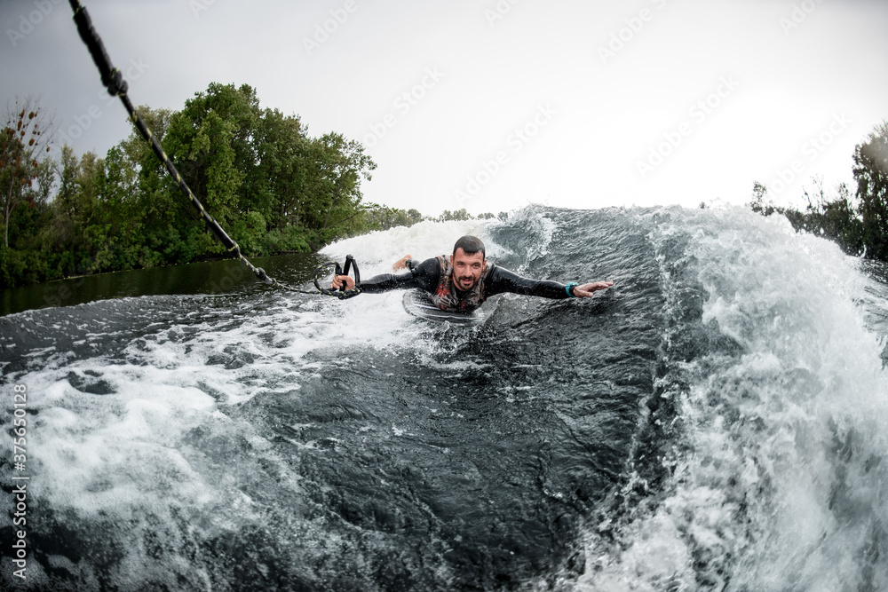 Obraz premium man holding rope and lying on wake surfboard rides on wave from motor boat