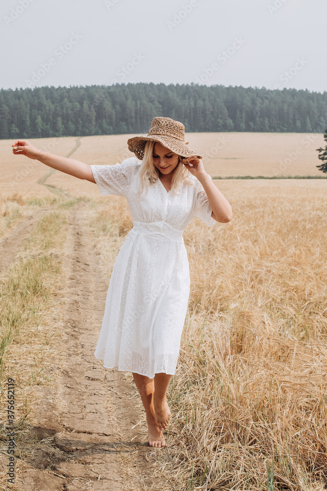 A beautiful girl in a white dress and a straw hat walks in the field. A blonde woman is resting in the village.
