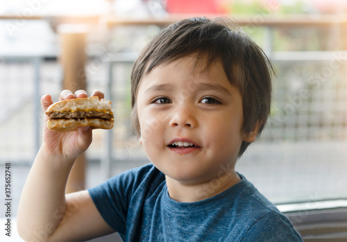 Preschool kid boy eats hamburger sitting in nursery cafe Cute happy boy holding hamburger and looking at camera with smiling face Child eating delicious homemade burger 