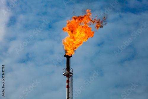 Burning orange gas torch on blue sky with clouds. Gas processing and oil refinery plant.