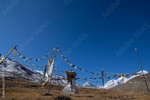 A Buddhist shrine surrounded with poles of colorful prayer flags on a windy clear day with a range of snow peaks in the background and blue sky.