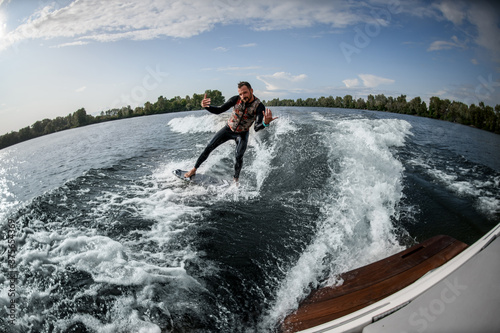 cheerful guy rides surfboard on the waves behind boat.