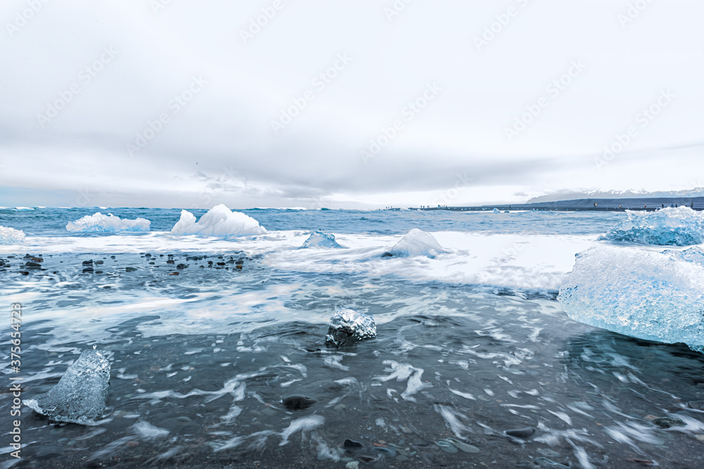 Waves shore long exposure motion on blue glacier iceberg ice floating on black sand by Jokulsaron lagoon lake diamond beach in Iceland