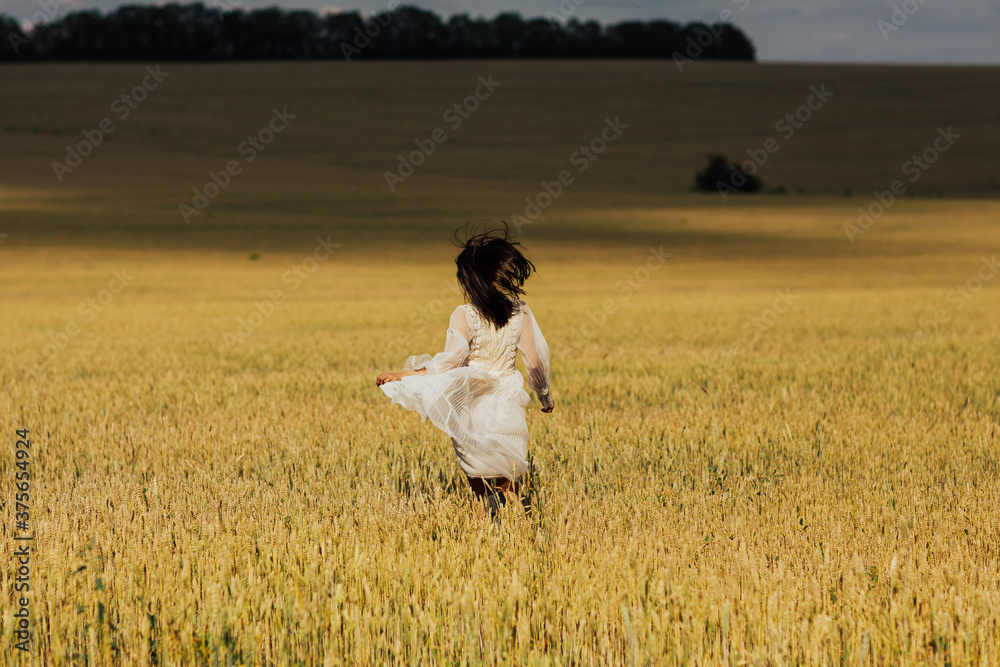 A beautiful cheerful girl runs along a wheat field in a beige dress. Back view. A beautiful cheerful girl runs along a wheat field in a beige dress. Back view.
