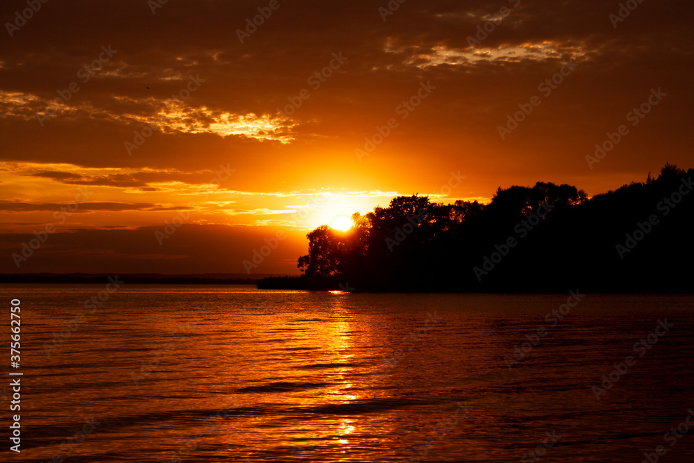 Horizontal landscape photography of an adorable warm orange summer sunset above the sea with dark silhouette of unknown island, calm water and dark clouds in the sky on the background