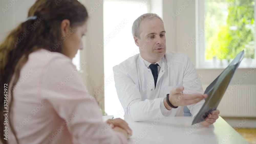 Portrait of confident doctor talking with patient showing lungs x-ray. Professional Caucasian man explaining radiograph to blurred young woman sitting at front. Pulmonologist consulting client. Portrait of confident doctor talking with patient showing lungs x-ray. Professional Caucasian man explaining radiograph to blurred young woman sitting at front. Pulmonologist consulting client.