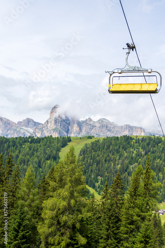 Yellow chairlift connecting valley floor to high peaks of Italian Alps. Sassongher Dolomite mountain is visible in the background, surrounded by green meadows. Colfosco - Val Badia, Italy