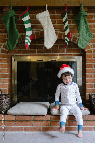 boy in pajamas and Santa hat sits in front of fireplace with stockings photo