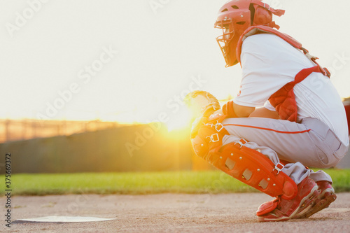 Baseball catcher squatting behind home plate backlit by sun.
