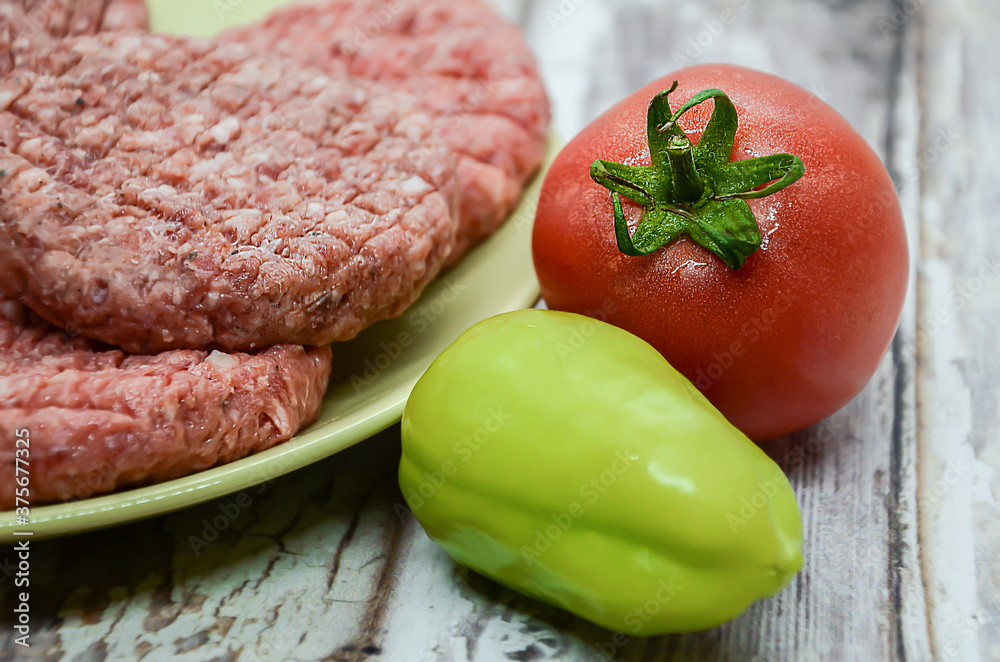 Ripe tomato and bell peppers on a background of raw homemade chopped beef burgers on a plate and a wooden table. BBQ preparation, beef products, traditional meat dish. Balanced diet. Ripe tomato and bell peppers on a background of raw homemade chopped beef burgers on a plate and a wooden table. BBQ preparation, beef products, traditional meat dish. Balanced diet.