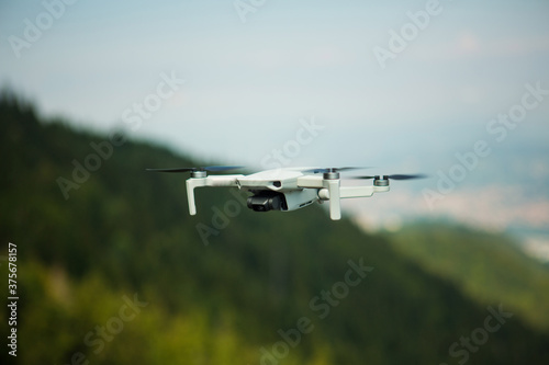 Mini white drone  in front of a hill in nature flying in a blue sky. The camera of the drone is facing towards focus. A flying drone captured from a high position.  A farest green backogrund with sky.