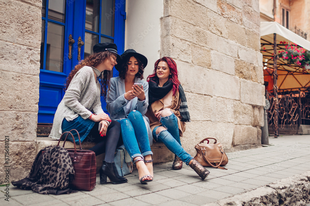 Three girlfriends using smartphone on city street. Stylish women sitting by cafe and having fun chatting outdoors Three girlfriends using smartphone on city street. Stylish women sitting by cafe and having fun chatting outdoors