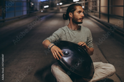 young bearded man playing handpan while sitting on the road, musical harmony in the middle of an evening urban city photo