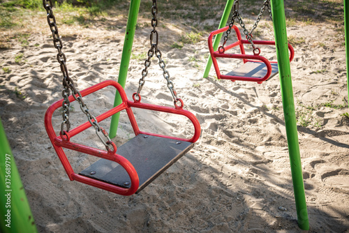 Empty swing-set on playground because of COVID lockdown. Sand underneath the swings and a grassy area, then many trees behind. Good summer camp image