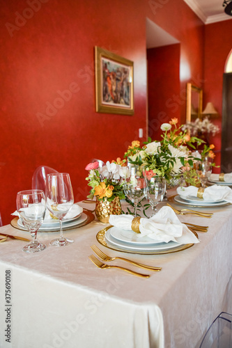 Elegant table setting with gold accents and red wall, clear chairs and flowers
