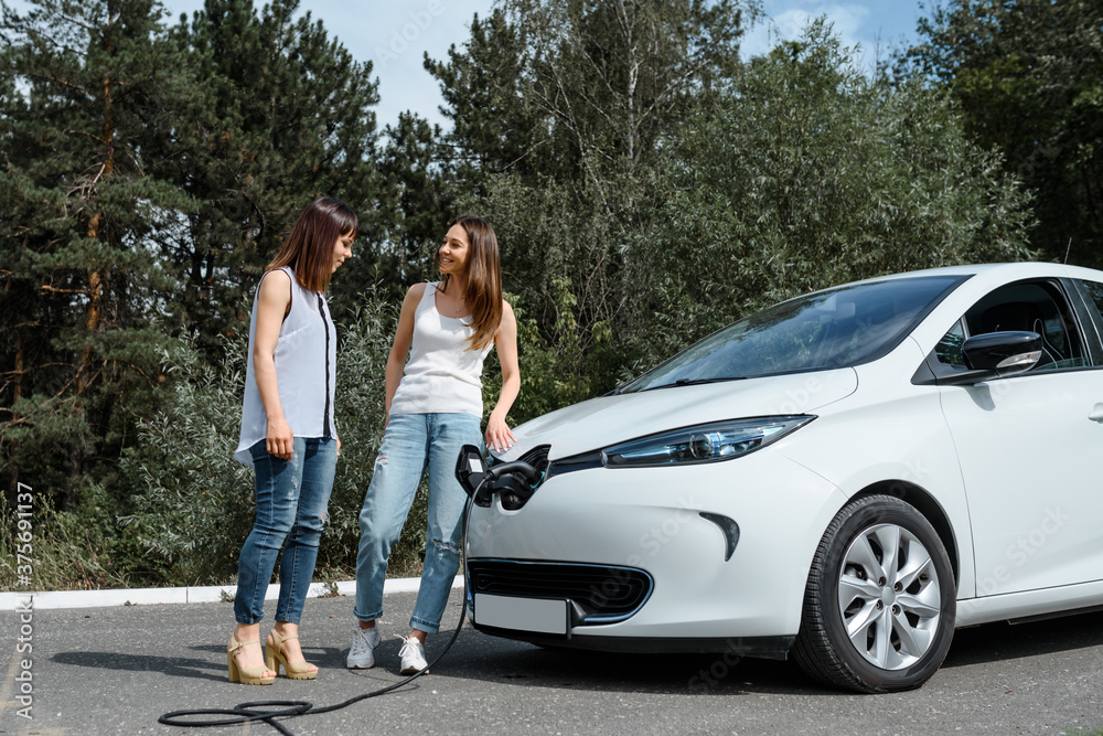 Two happy young beautiful women are talking to each other until their electric car is charging at the charging station situated in the forest. Two happy young beautiful women are talking to each other until their electric car is charging at the charging station situated in the forest.