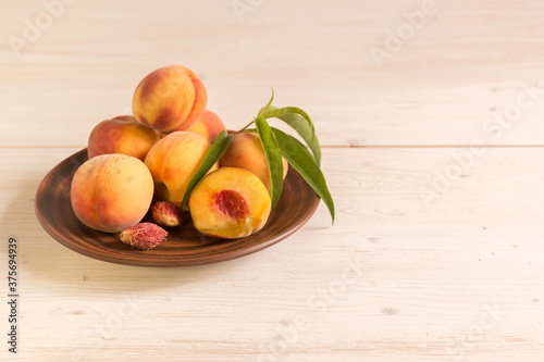 Many ripe velvety large and beautiful peaches with leaves lie in a plate on a white wooden table. Sunny
