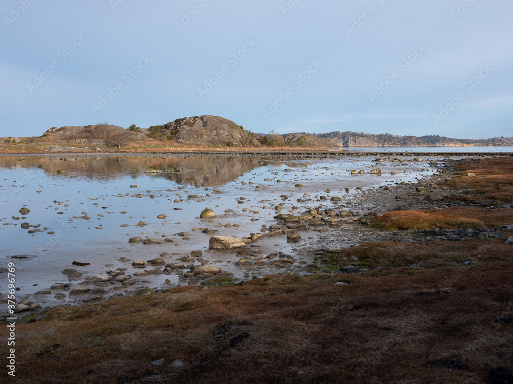 Beautiful photo of a coastline. It is shot in early morning at low tide, exposing hundreds of rocks lying on the sea bed. It is early spring and frost in the grass in the foreground. 